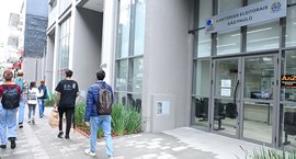 A fotografia mostra a fachada do edifício com amplas janelas e portas duplas de vidro, com a pla...