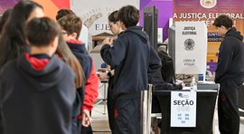 Fotografia colorida, em plano médio e ambiente interno, mostrando estudantes em uma fila para pa...