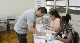 Duas mesárias e um eleitor assinando folha em Mesa Receptora de Votos