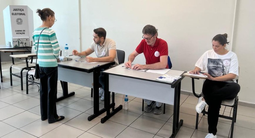 A imagem mostra o interior de uma seção eleitoral. Uma eleitora está de pé diante de uma mesa on...