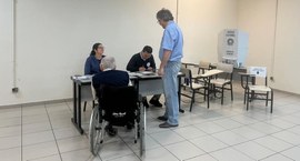 Fotografia em ambiente interno de uma seção eleitoral. Em primeiro plano, um idoso em uma cadeir...