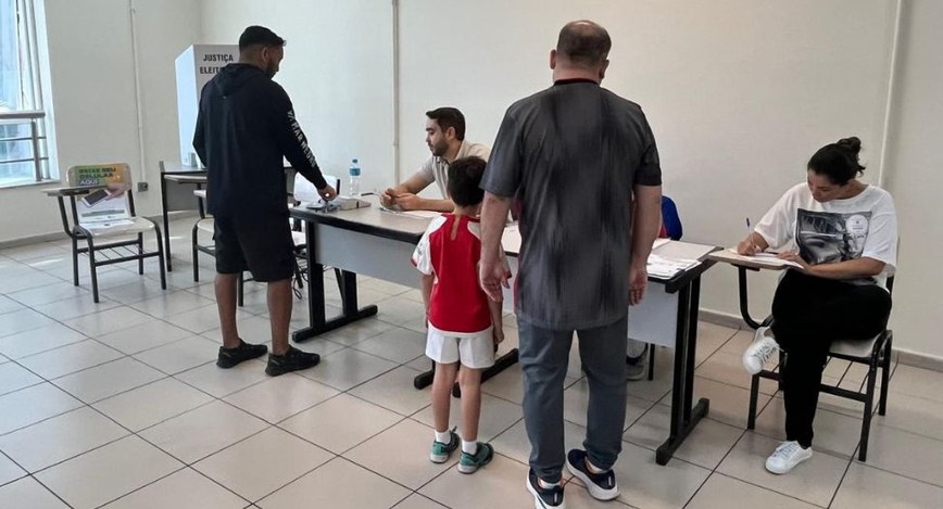 Fotografia colorida em plano médio mostrando o interior de uma seção eleitoral. Eleitores, inclu...