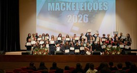 Fotografia de um grupo de estudantes e adultos reunidos no palco de um auditório. Eles posam sor...