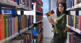 Servidora segura livro posicionada entre as estantes da biblioteca do TRE-SP