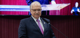 Fotografia que retrata, em primeiro plano, um homem de pele clara, cabelos grisalhos e bigode, u...