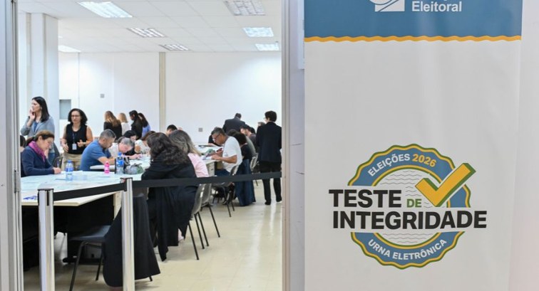 No primeiro plano, em foco, está um banner branco com o selo do Teste de Integridade das Eleiçõe...
