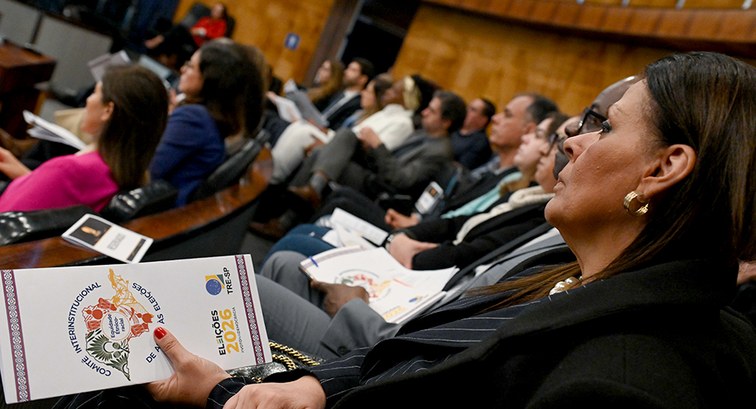 Mulher em plateia segura folder sobre o Comitê Interinstitucional de Apoio às Eleições 2026 do T...