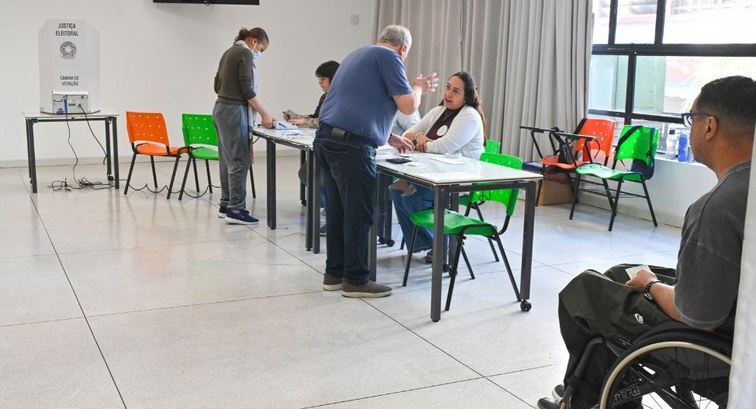 Seção eleitoral com cabine de votação ao fundo da sala, e uma pessoa em cadeira de rodas esperan...
