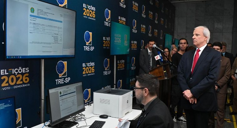 Na sala de imprensa do TRE-SP, com um banner azul escuro com o logo das Eleições 2026 ao fundo, ...