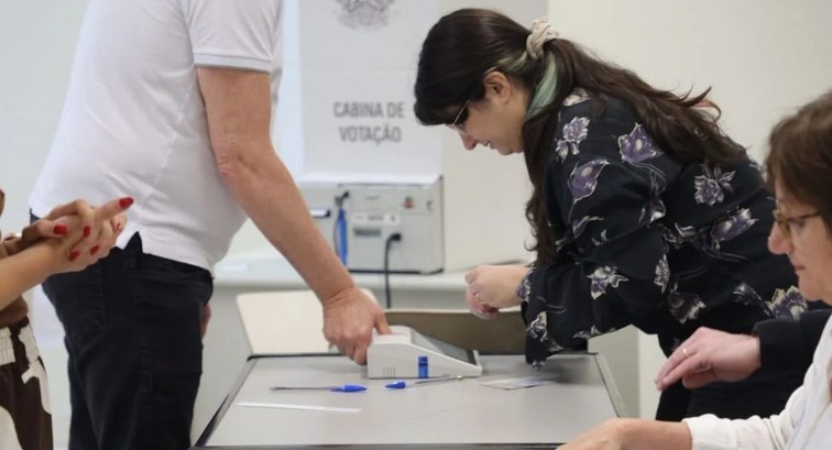 Mesa de seção eleitoral, com uma mesária procurando nome de eleitor em caderno de votação e outr...