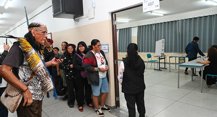 Uma fila de eleitores indígenas esperam ao lado da porta de uma sala, que funciona como seção el...