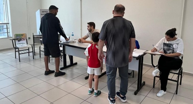 Fotografia colorida em plano médio mostrando o interior de uma seção eleitoral. Eleitores, inclu...
