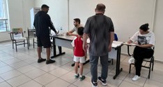 Fotografia colorida em plano médio mostrando o interior de uma seção eleitoral. Eleitores, inclu...