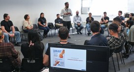 Fotografia  mostrando uma dinâmica em grupo ocorrendo dentro de uma sala de reuniões. As pessoas...