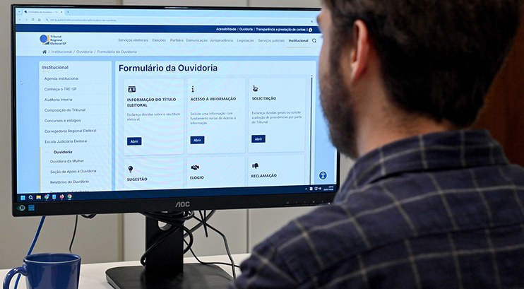Pessoa olha tela de um computador em que aparece aparece a foto da página da Ouvidoria do TRE-SP