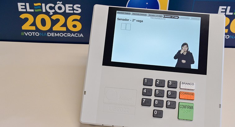 Foto focando na tela de uma urna 2022, posicionada em cima de uma mesa de cor clara. A pessoa es...