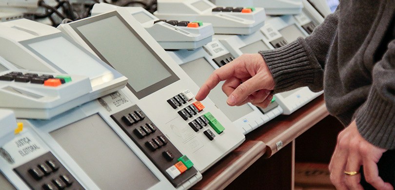 Urnas do modelo 2020 também foram testadas. Fotografia da mão de uma pessoa testando uma urna. Outras diversas urnas estão enfileiradas ao l...