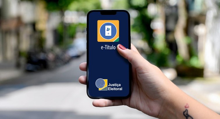Uma mão segura um smartphone em primeiro plano, exibindo na tela azul-escura o aplicativo do e-T...