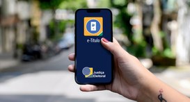 Uma mão segura um smartphone em primeiro plano, exibindo na tela azul-escura o aplicativo do e-T...