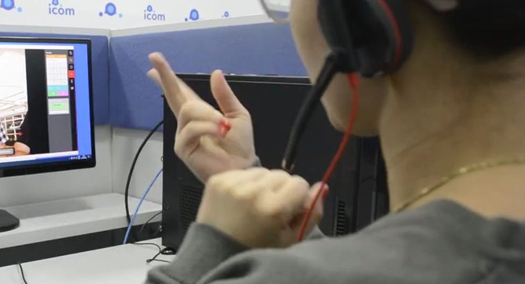 Visão por trás do ombro de uma pessoa de blusa cinza e usando headset, gesticulando em língua de...