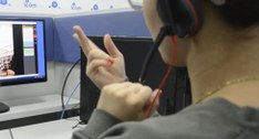 Visão por trás do ombro de uma pessoa de blusa cinza e usando headset, gesticulando em língua de...