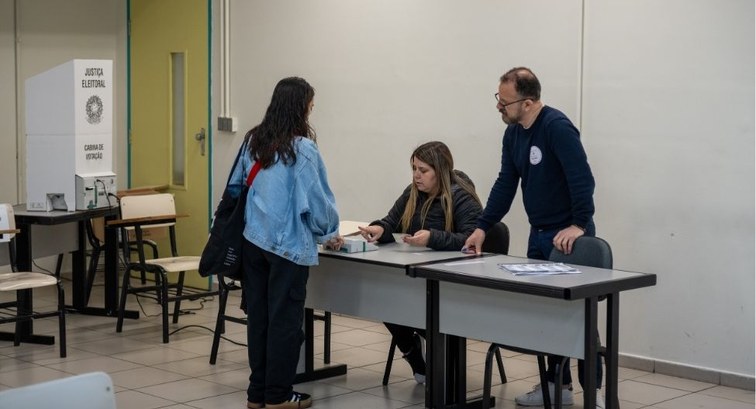 Uma eleitora de jaqueta jeans está em pé em um local de votação, entregando seu documento a dois...