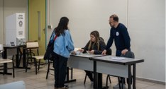 Uma eleitora de jaqueta jeans está em pé em um local de votação, entregando seu documento a dois...