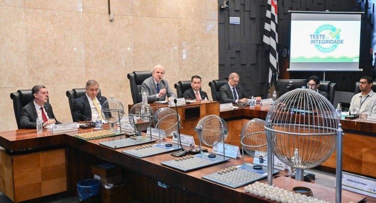 Fotografia em ambiente institucional com autoridades sentadas ao redor de uma grande mesa de mad...
