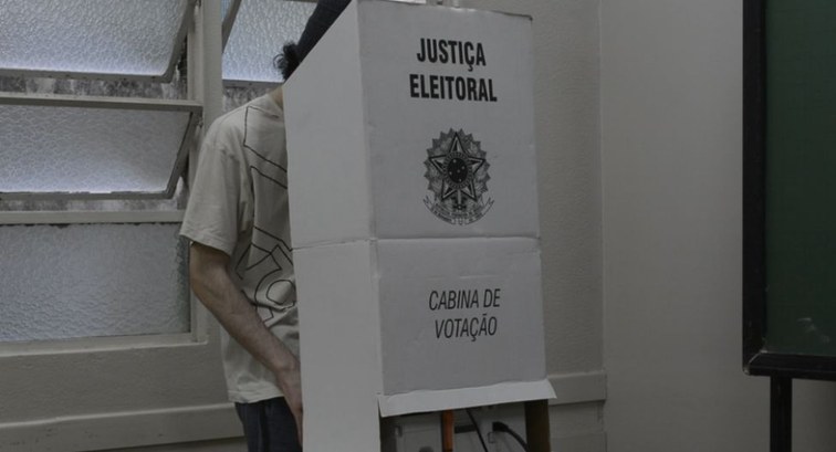 Pessoa votando atrás de uma cabina de papelão branca da Justiça Eleitoral brasileira, com o Bras...