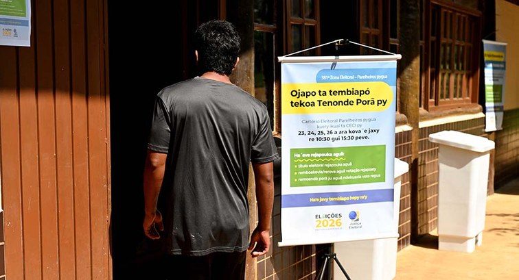 Homem de costas a entrar num prédio de madeira. Ao lado da entrada, há um cartaz da Justiça Elei...