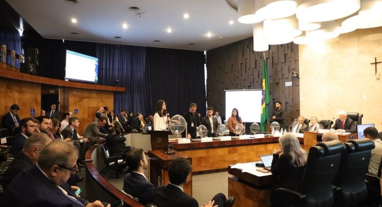 Em um auditório com formato de plenário, uma mulher fala ao microfone diante de uma bancada de m...