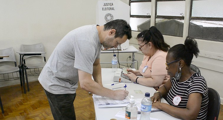Duas mesárias e um eleitor assinando folha em Mesa Receptora de Votos