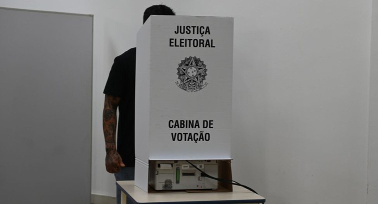 Pessoa de camiseta preta vota em urna eletrônica protegida por uma cabina branca da Justiça Elei...