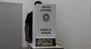 Pessoa de camiseta preta vota em urna eletrônica protegida por uma cabina branca da Justiça Elei...