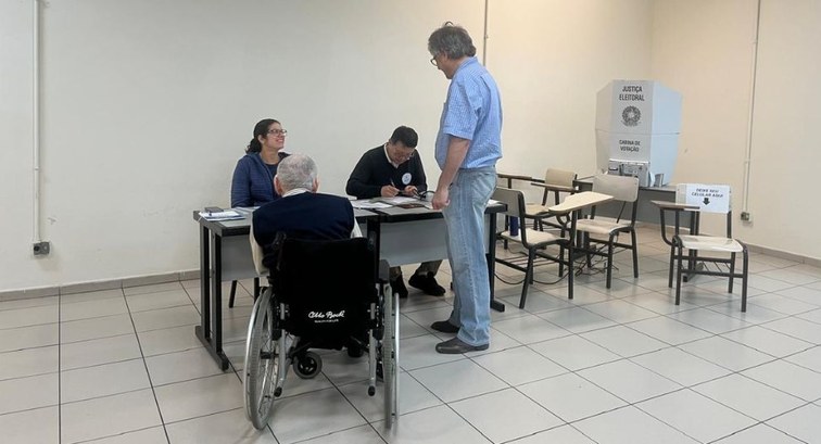 Fotografia em ambiente interno de uma seção eleitoral. Em primeiro plano, um idoso em uma cadeir...