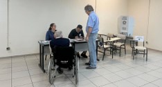 Fotografia em ambiente interno de uma seção eleitoral. Em primeiro plano, um idoso em uma cadeir...