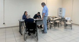 Fotografia em ambiente interno de uma seção eleitoral. Em primeiro plano, um idoso em uma cadeir...