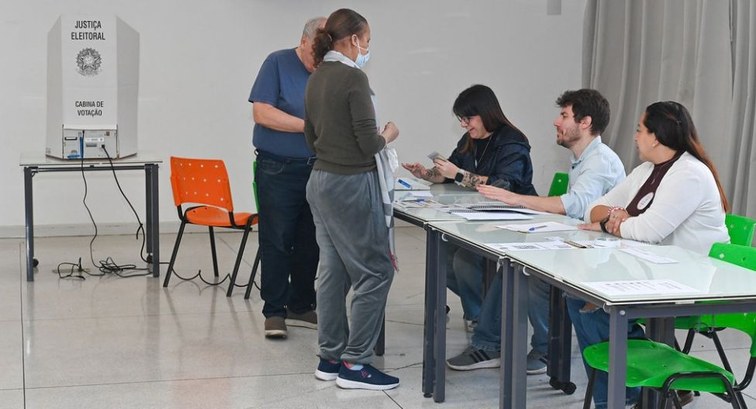 Fotografia em ambiente interno de uma seção eleitoral onde eleitores interagem com mesários sent...