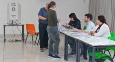 Fotografia em ambiente interno de uma seção eleitoral onde eleitores interagem com mesários sent...