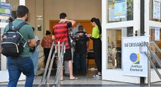 Fotografia em plano médio mostra eleitores na entrada de um local de votação, onde uma assistent...
