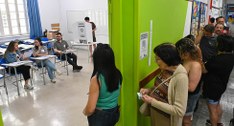 Seção eleitoral movimentada dentro de uma sala de aula, onde mesários atendem eleitores sentados...