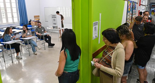 Seção eleitoral movimentada dentro de uma sala de aula, onde mesários atendem eleitores sentados...