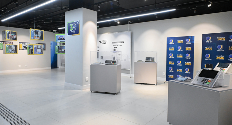 Espaço expositivo iluminado com urnas eletrônicas de diferentes gerações sobre mesas. Ao fundo, ...