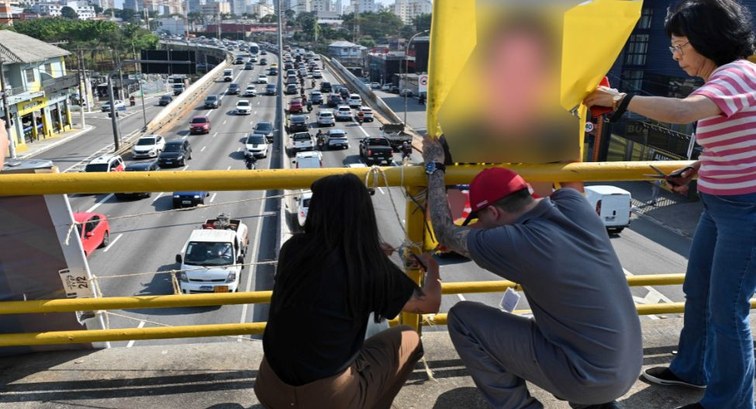 Em primeiro plano, três pessoas retiram um cartaz amarelo na grade de proteção de uma passarela ...