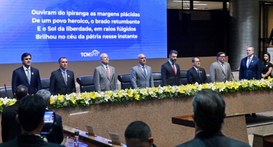Fotografia de um evento formal em auditório com autoridades atrás de uma bancada decorada com fl...