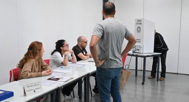 Três mesários estão sentados em uma mesa branca com cadernos de votação, observando um homem em ...