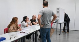 Três mesários estão sentados em uma mesa branca com cadernos de votação, observando um homem em ...
