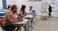 Fotografia em plano médio de uma seção eleitoral em sala de aula. Três mesários estão sentados e...