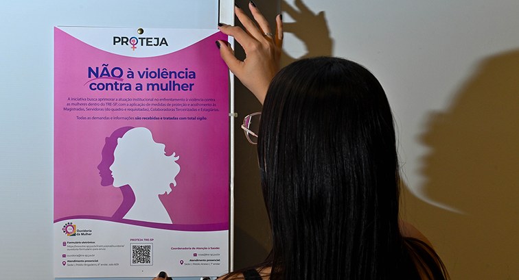 Uma pessoa vista de costas e de perfil segura um cartaz rosa na parede com a mensagem "NÃO à vio...