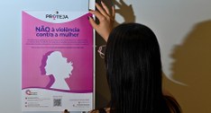 Uma pessoa vista de costas e de perfil segura um cartaz rosa na parede com a mensagem "NÃO à vio...